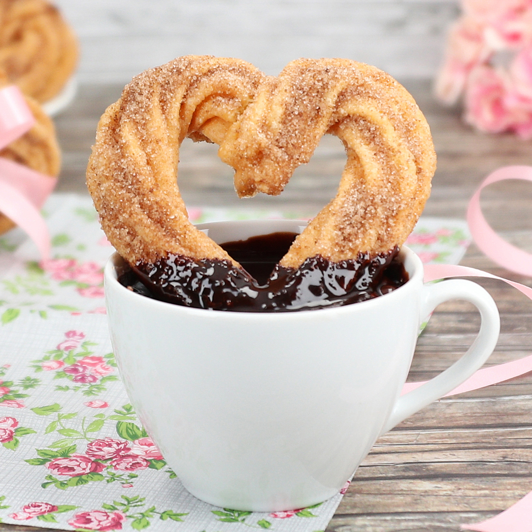 Das Bild zeigt Churro in Herzform, in einer Tasse mit Schokoladensauce