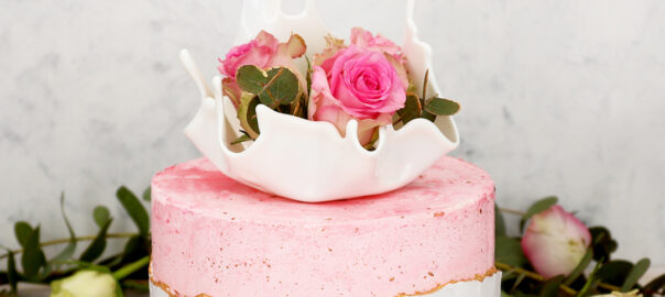 Das Bild zeigt eine rosa und weiße Torte mit einer goldenen Linie auf der ein Cake Topper liegt in dem Rosen sind