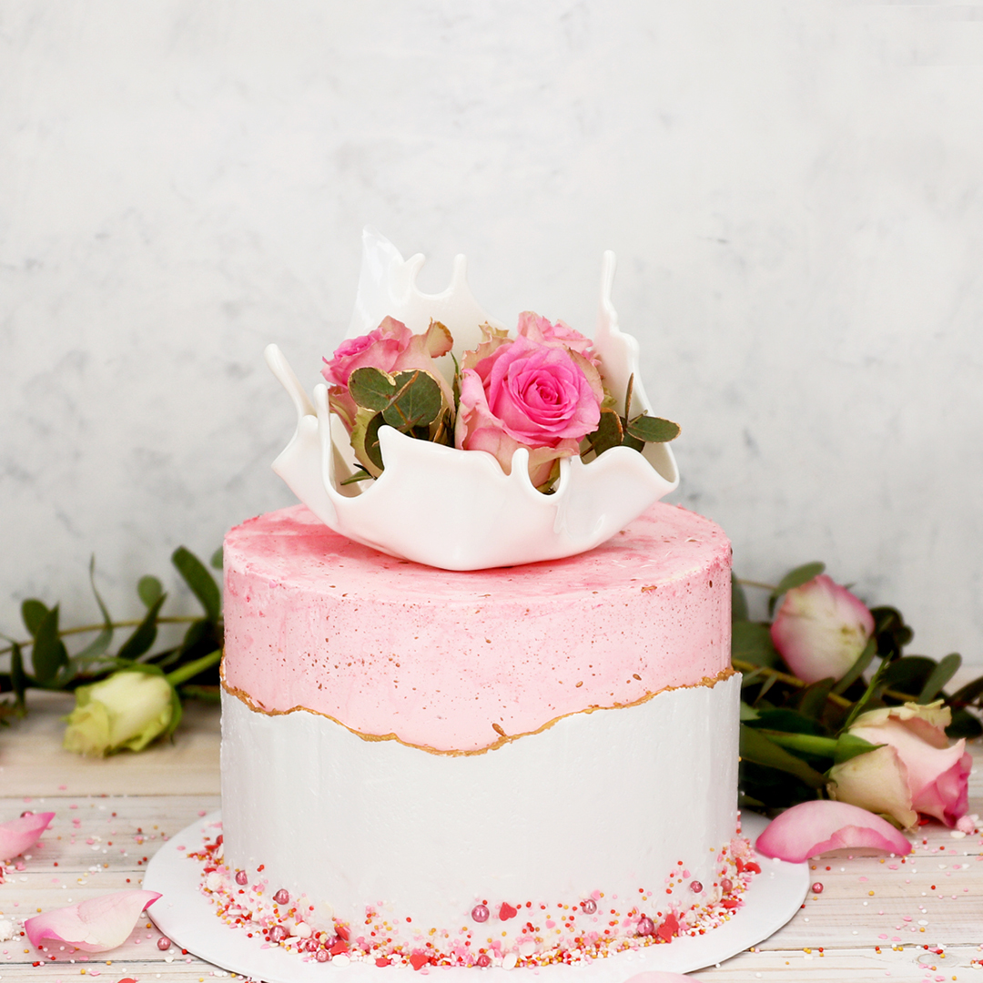 Das Bild zeigt eine rosa und weiße Torte mit einer goldenen Linie auf der ein Cake Topper liegt in dem Rosen sind