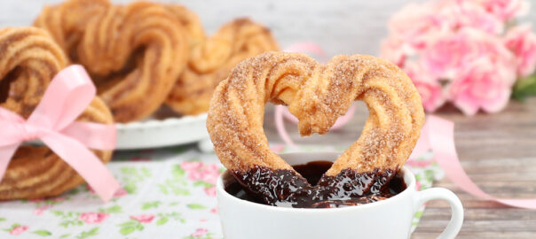 Das Bild zeigt Churros in Herzform in einer Tasse mit Schokosauce