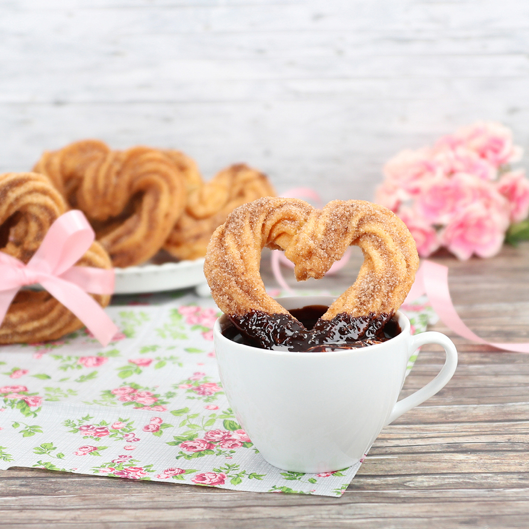 Das Bild zeigt Churros in Herzform in einer Tasse mit Schokosauce